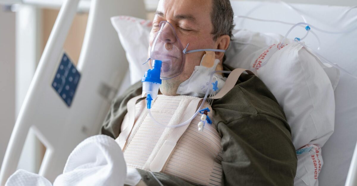 An elderly man lies in a hospital bed wearing an oxygen mask for medical care