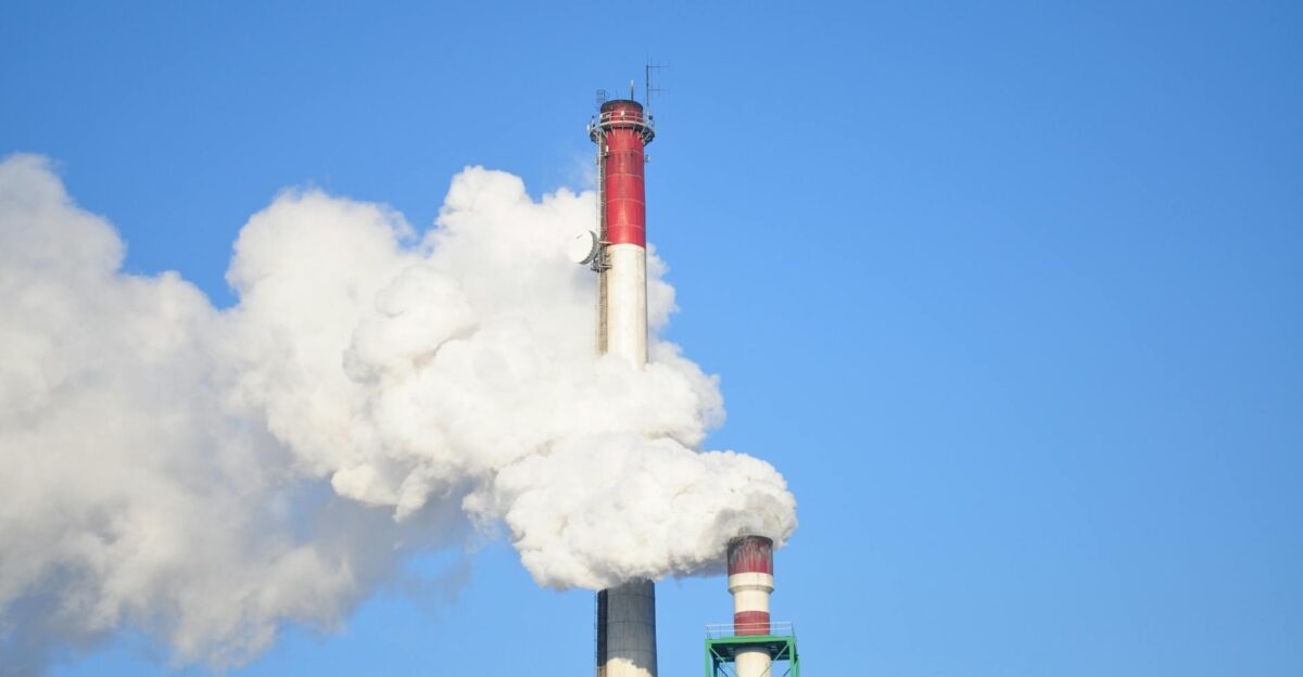 A tall industrial smokestack releasing thick white smoke against a clear blue sky