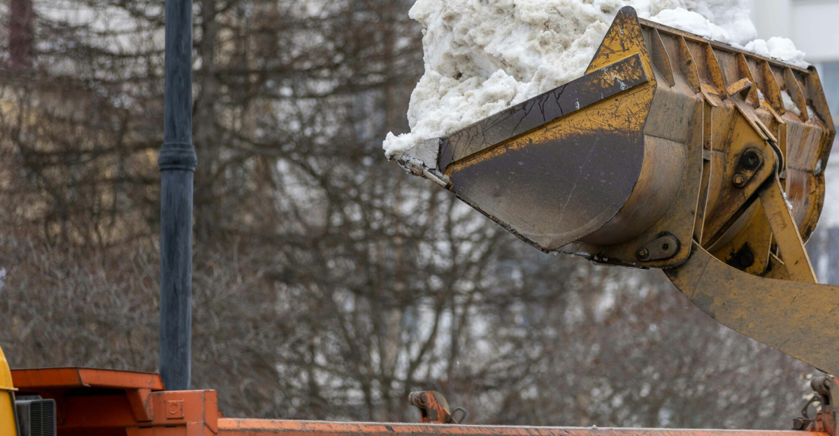 Excavator clearing snow into dump truck on city street during winter.