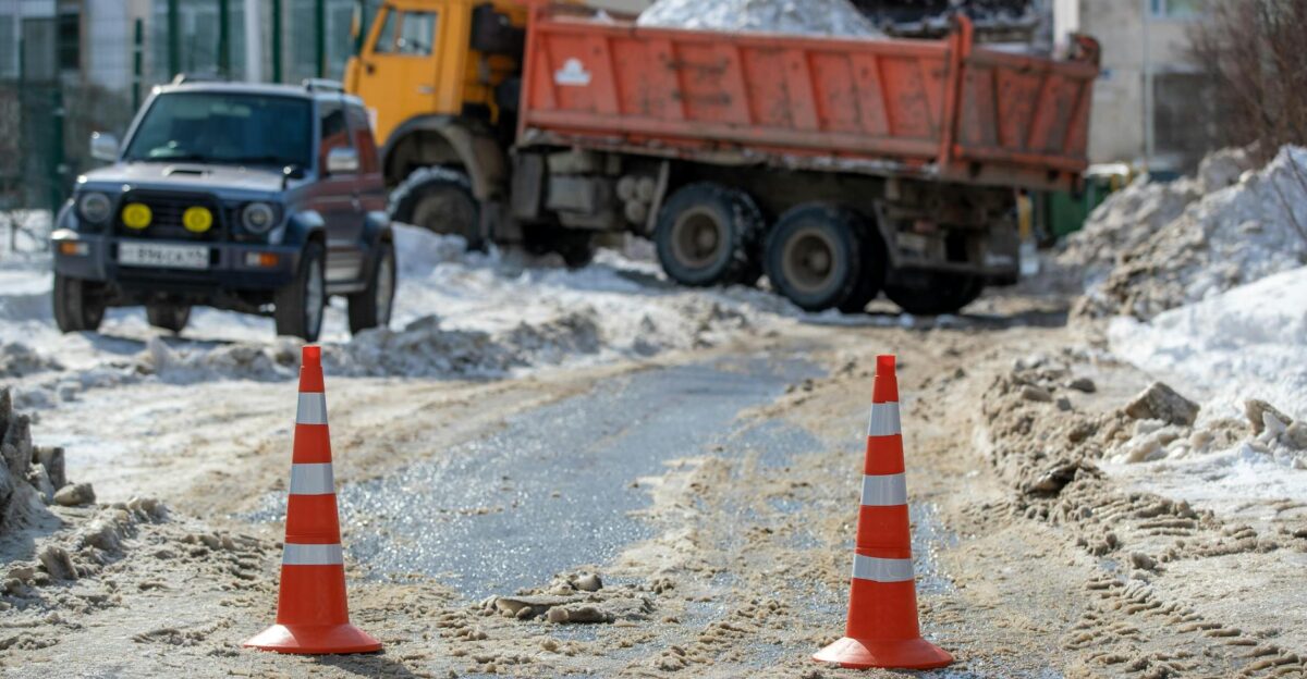 Orange traffic cones on a snowy road with parked vehicles and construction equipment in the background