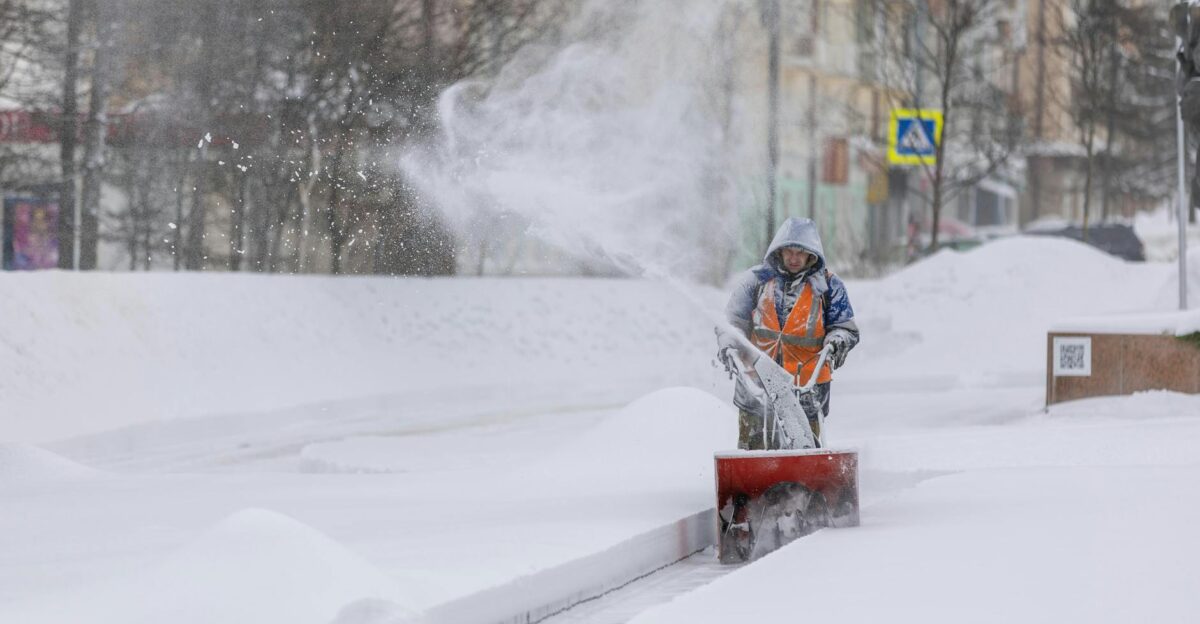 Person using a snow blower to clear a city sidewalk during a heavy winter snowfall