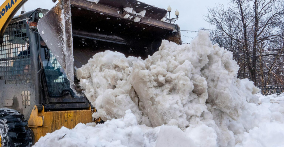 A yellow tractor clears snow on a city street after a winter snowstorm, showcasing urban cleanup.