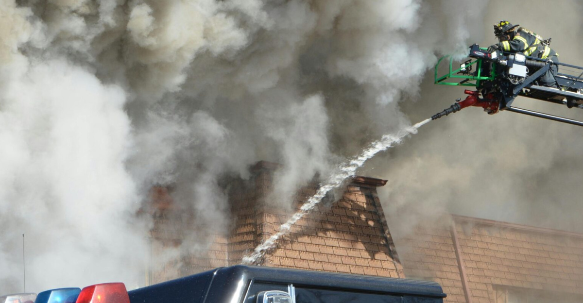 Firefighters on a ladder extinguishing a huge building fire, emitting thick clouds of smoke into the sky.