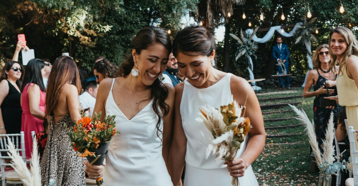 Two brides happily walk down the aisle at an outdoor wedding ceremony surrounded by friends and family