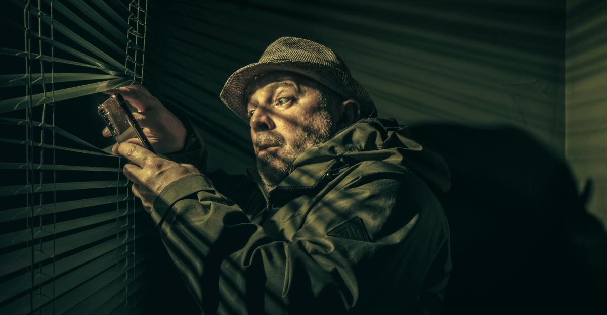 A man in a hat suspiciously peering through blinds holding a phone in shadowy light