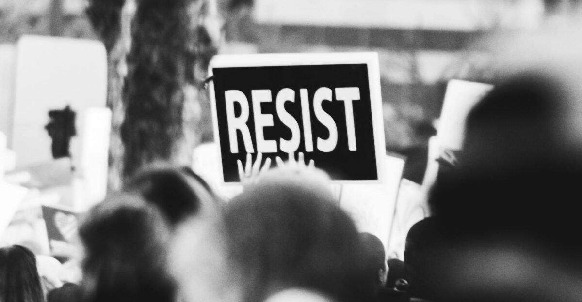Monochrome image showing a crowd holding a Resist sign at a protest
