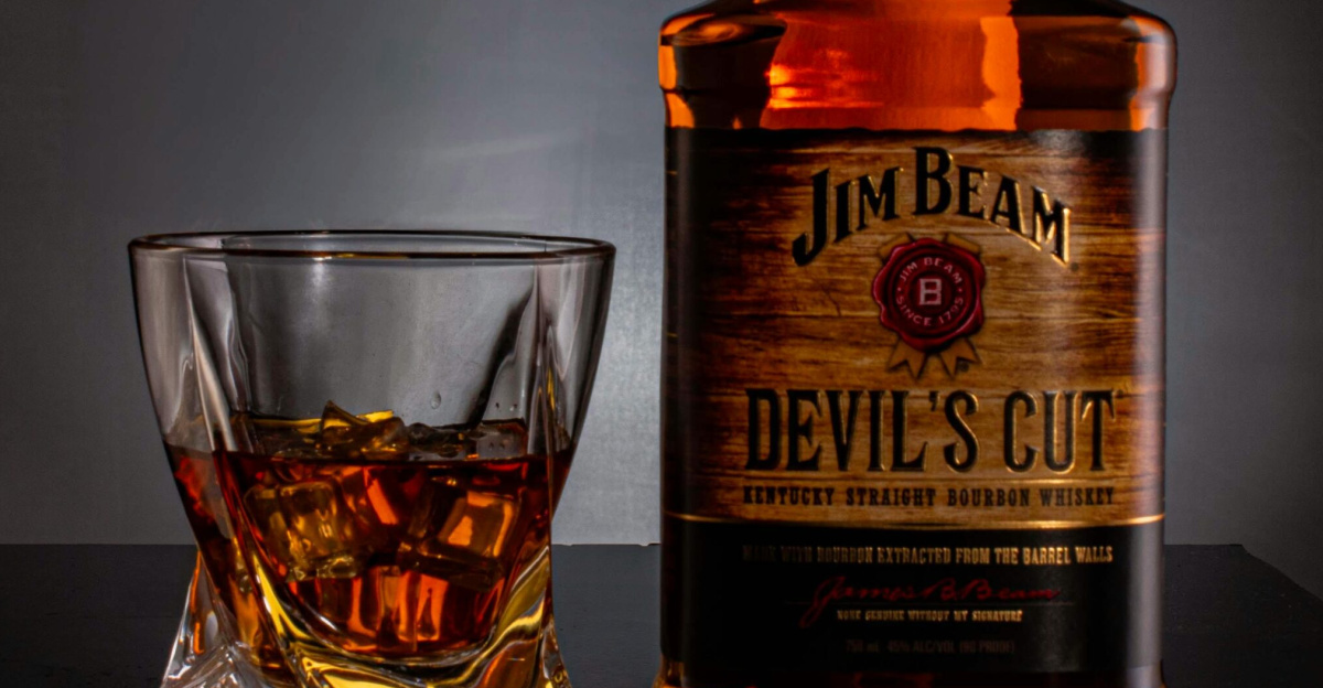 Bottle of Jim Beam Devil's Cut Bourbon and a glass filled with whiskey on a dark background.