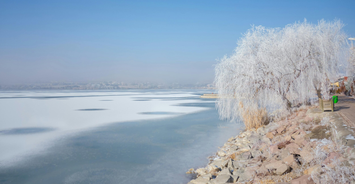 A serene winter landscape of a frozen lake with frost-covered trees under a clear blue sky.