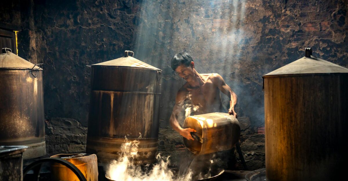 A man works in a traditional distillery surrounded by steam and sunlight beams