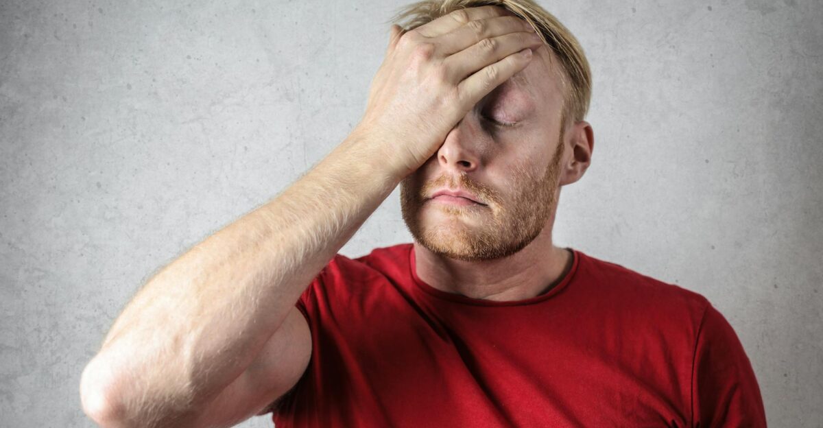 A frustrated man in a red shirt holds his head in stress against a neutral background