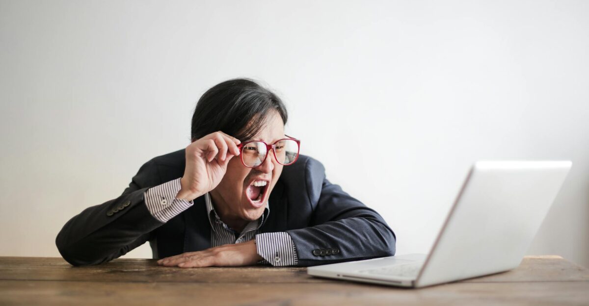 Modern Asian man in jacket and glasses looking at laptop and screaming with mouth wide opened on white background