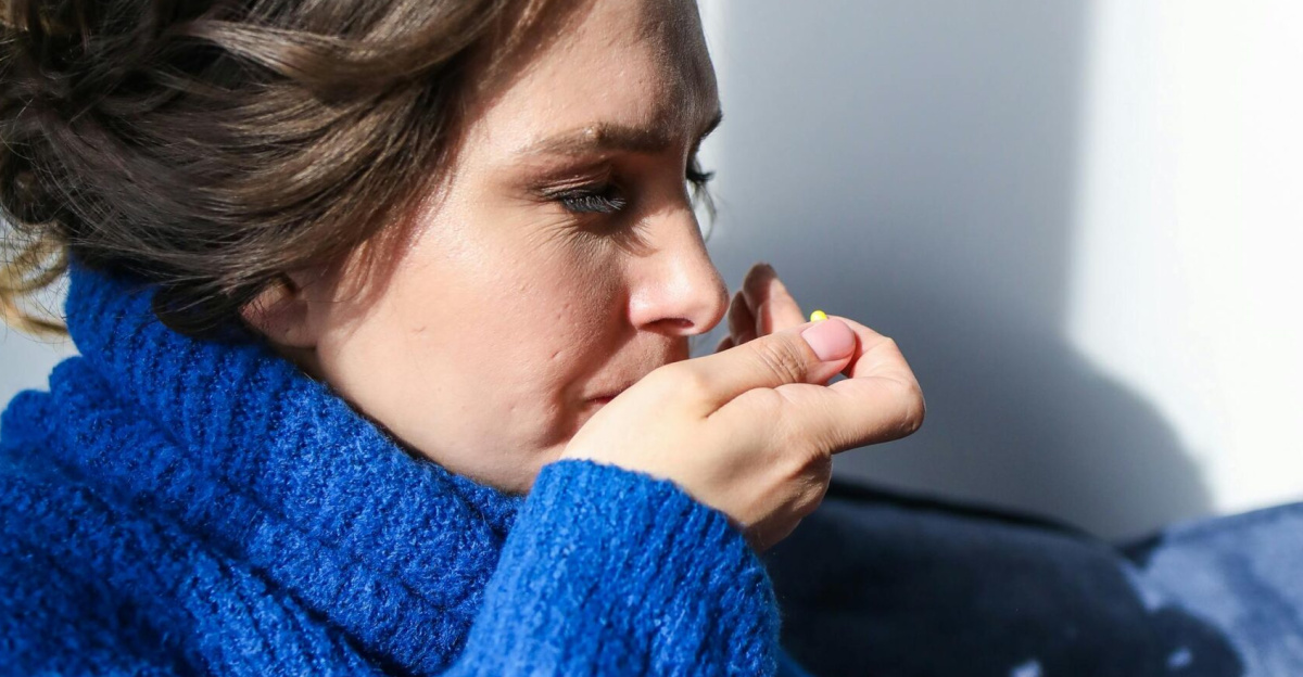 A woman in a blue sweater taking medication with a hot drink, showing flu symptoms.