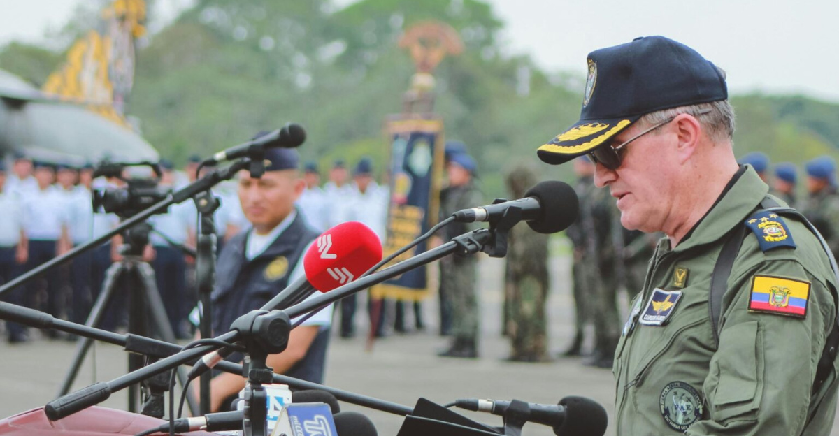 A military officer delivers a speech at an outdoor ceremony with an aircraft and soldiers in the background.