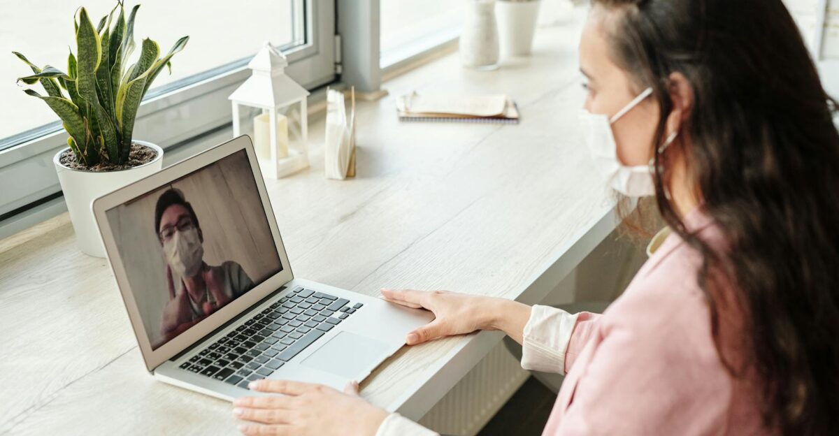 Woman and doctor video calling on laptop both wearing masks symbolizing telehealth during COVID-19