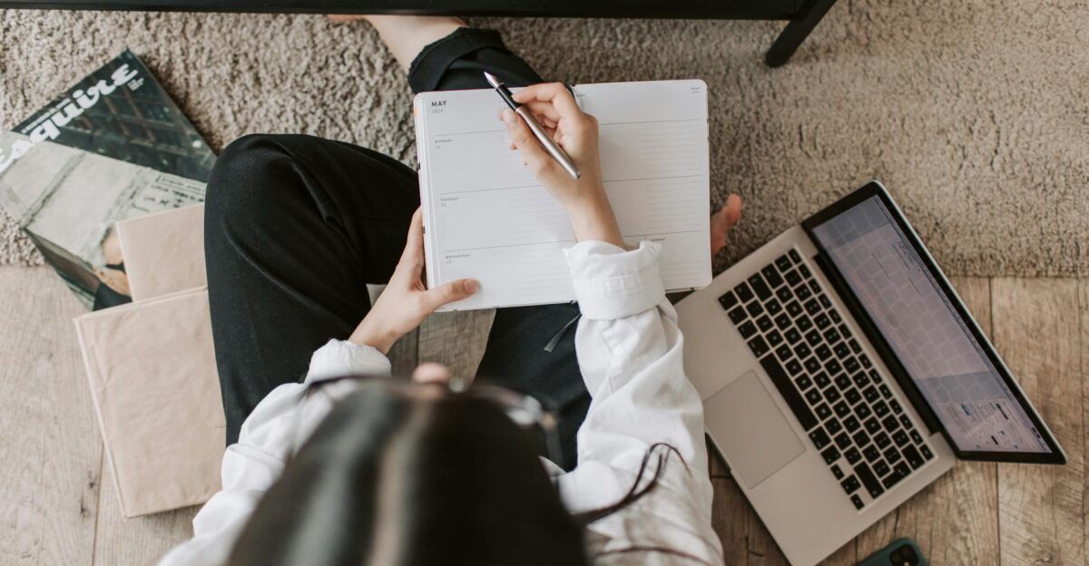 Top view of anonymous woman in casual wear sitting on floor with laptop and smartphone and creating plan on notebook while resting during break in modern living room