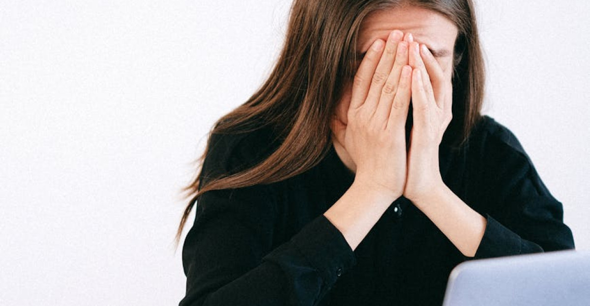 A woman covers her face with hands in stress sitting at an office with a laptop and papers