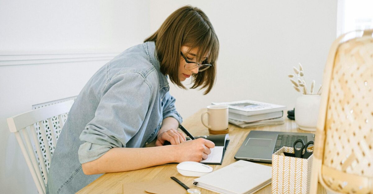 A young woman working from home writing notes at a desk with a laptop and papers