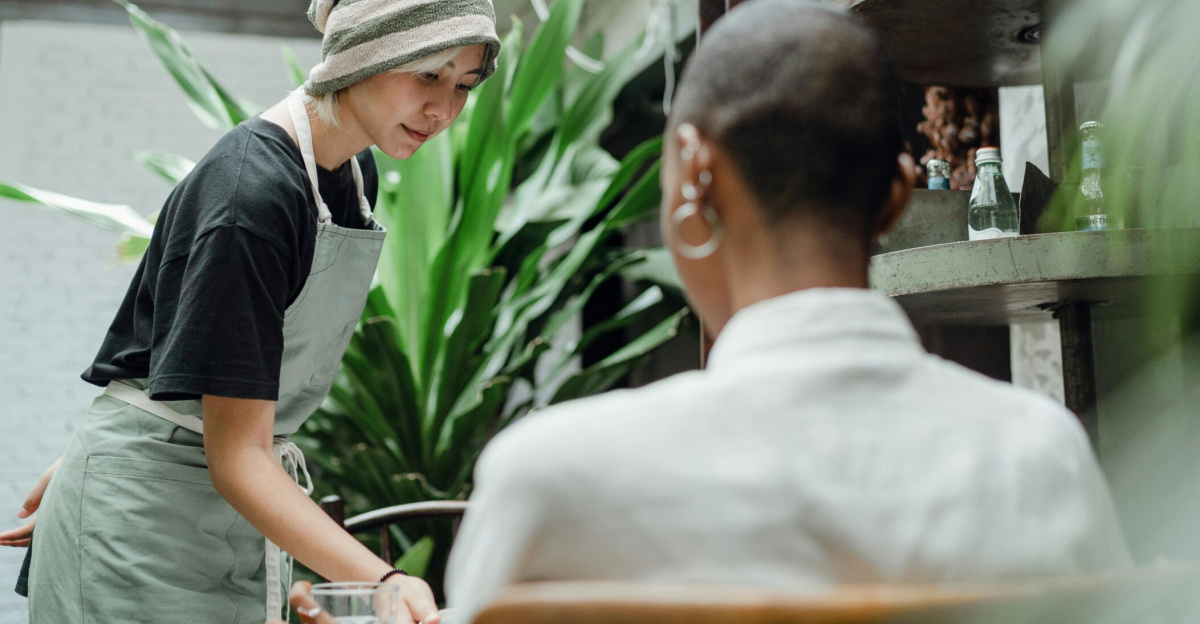 Positive young Asian waitress in uniform smiling and serving delicious food to female customer in modern cozy restaurant full of lush plants