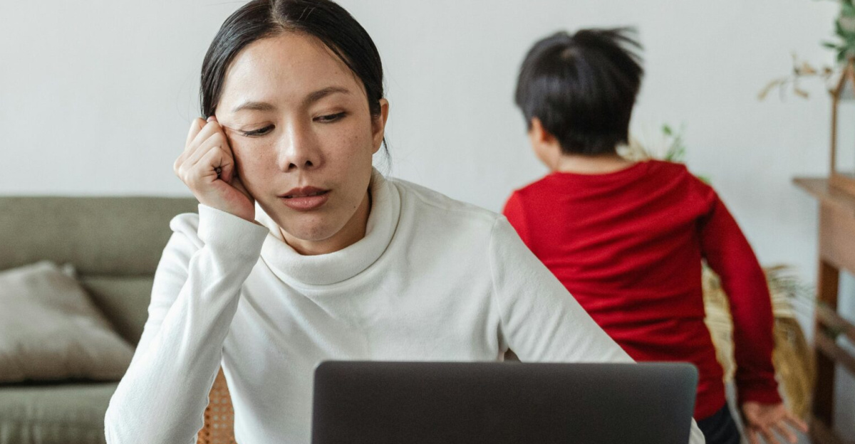 A tired mother working on a laptop while her children play around indoors, highlighting remote work challenges.