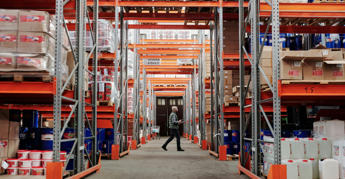 A man walking through a large industrial warehouse with stacked shelves filled with goods and products.