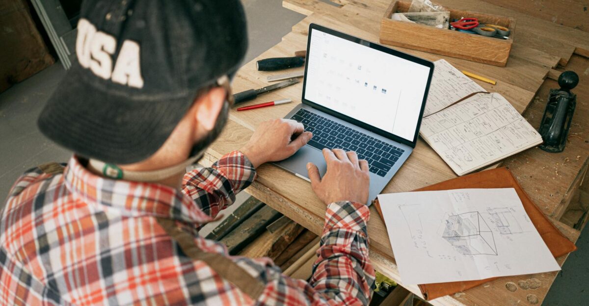 A woodworker in a workshop uses a laptop for furniture design planning and project management
