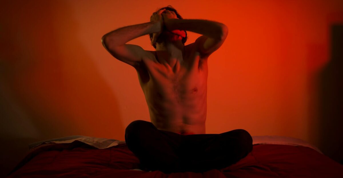 A topless man showing emotional distress sitting on a bed in a dimly lit room