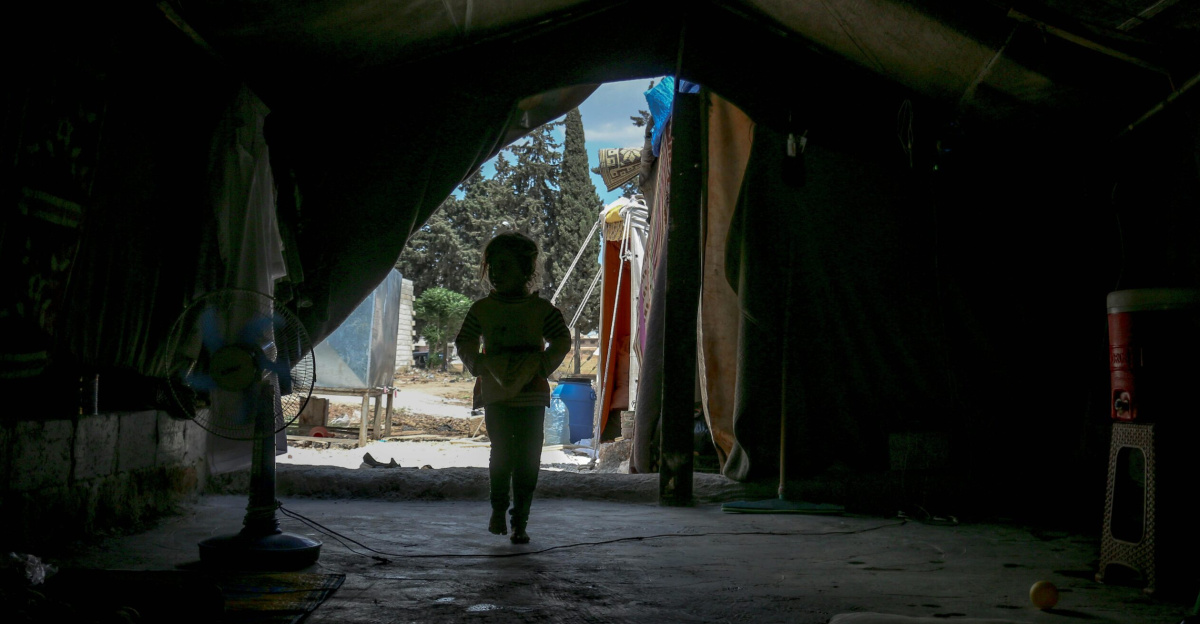 A silhouette of a child standing inside a refugee tent facing an outdoor area.