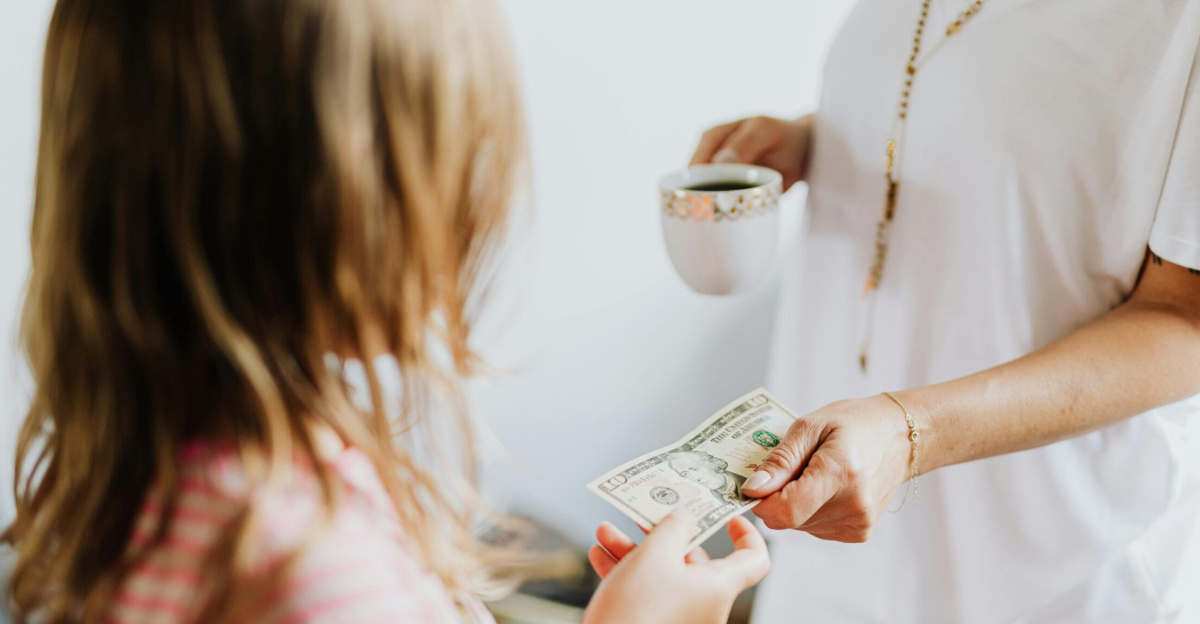 A woman hands a dollar bill to her daughter while holding a coffee cup, indoors.