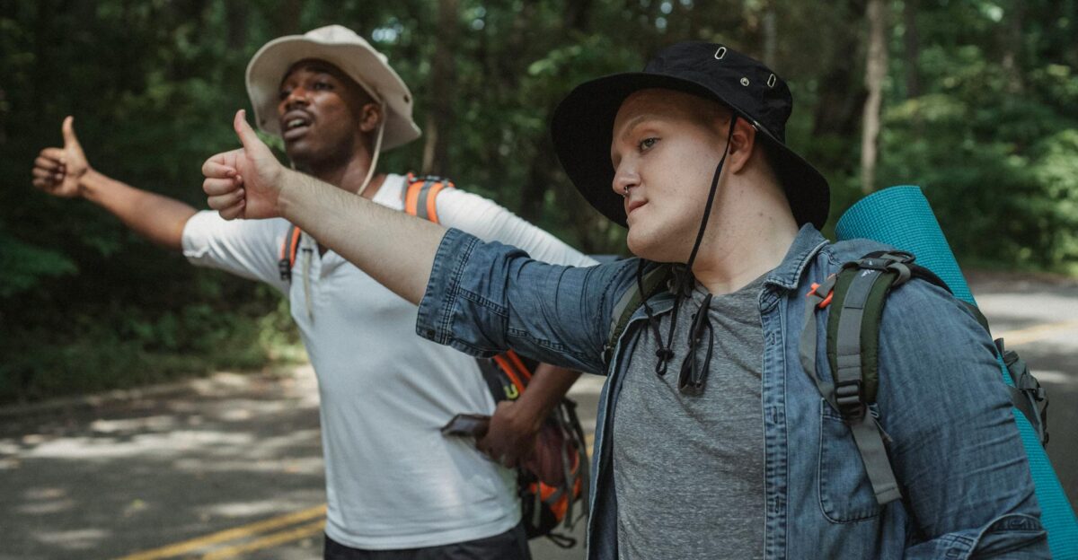 Young multiracial male backpackers in panama hats with hiking equipment standing with thumbs up on roadway against forest and looking away