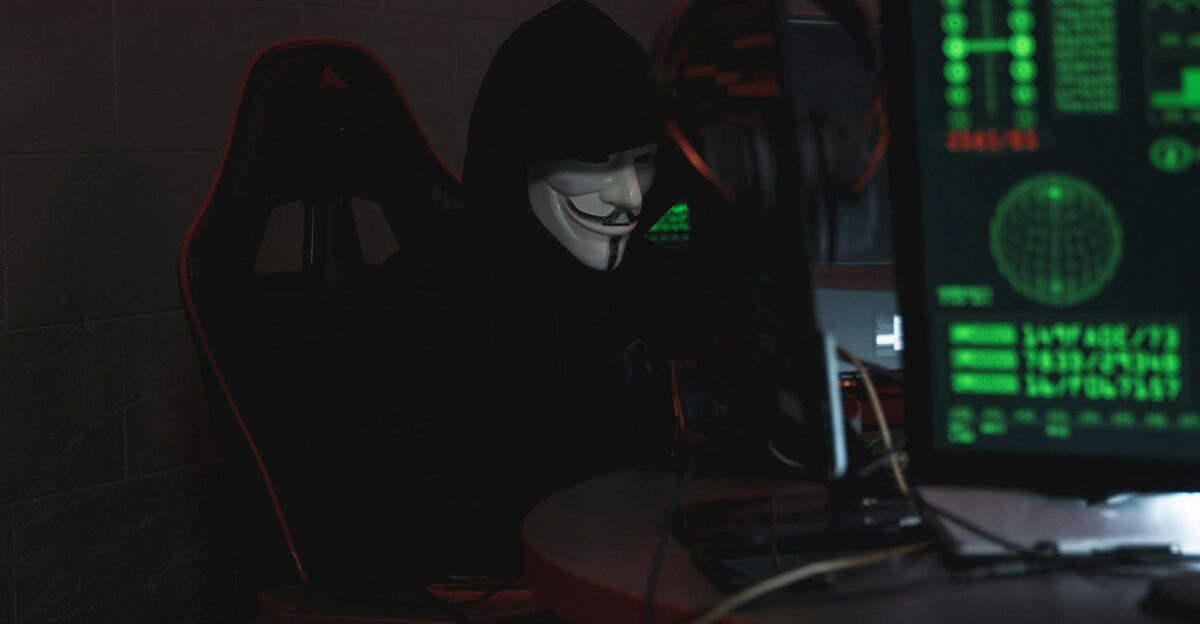 An anonymous hacker wearing a Guy Fawkes mask sits at a computer in a dimly lit room engaged in cyber activities