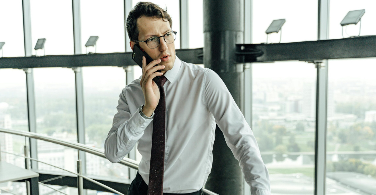 Young professional in a white shirt and tie making a phone call in a high-rise office with city view.