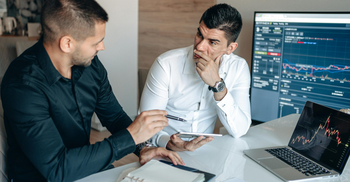 Two businessmen discussing stock market trends with trading screens in an office setting.