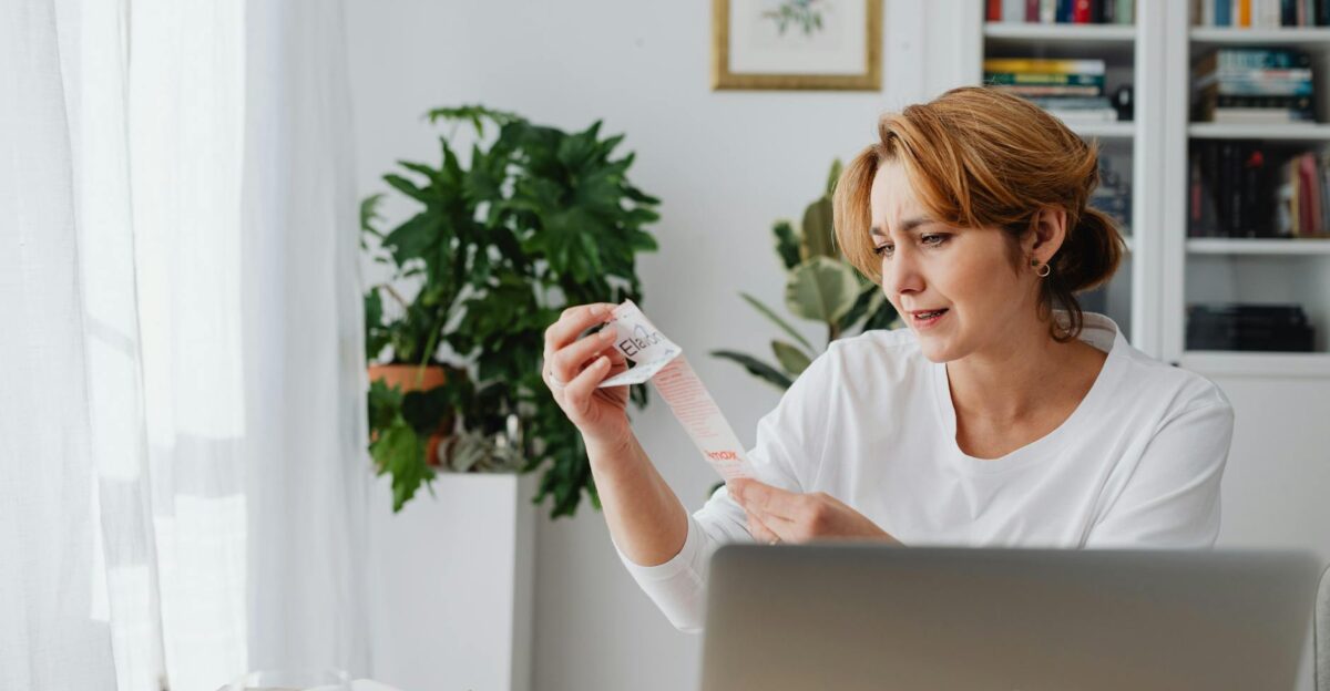A woman in a white shirt sits indoors examining a lengthy receipt with a concerned expression