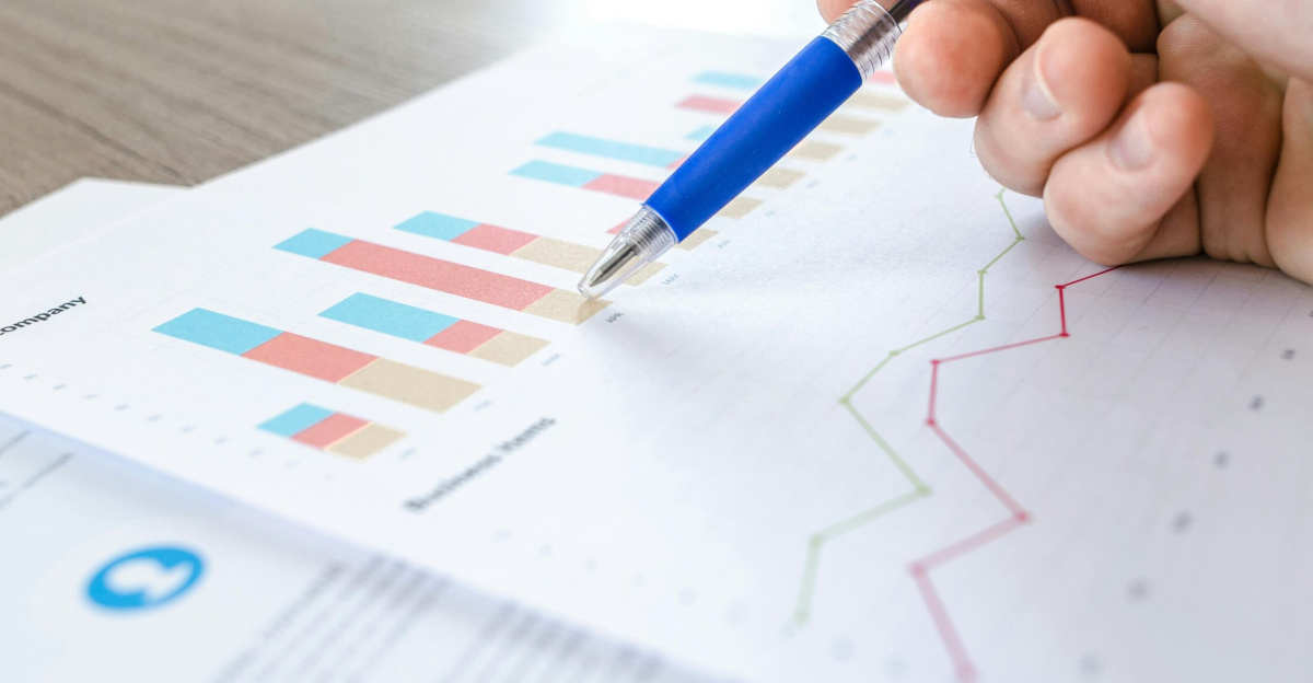 A close-up of a hand with a pen analyzing data on colorful bar and line charts on paper.