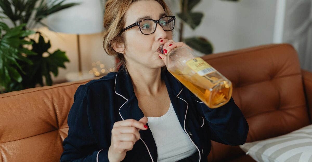 A woman in pajamas sits on a couch drinking from a glass bottle indoors