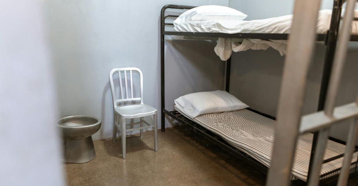 A stark prison cell featuring a bunk bed metal chair and toilet