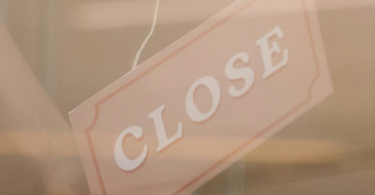 Close-up of a hand holding a 'Closed' sign through a glass door, reflection visible.