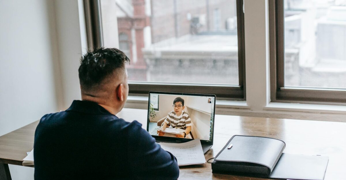 Back view of male in casual clothes sitting at table on chair while having video chat on laptop with pupil near papers and briefcase in light apartment near window