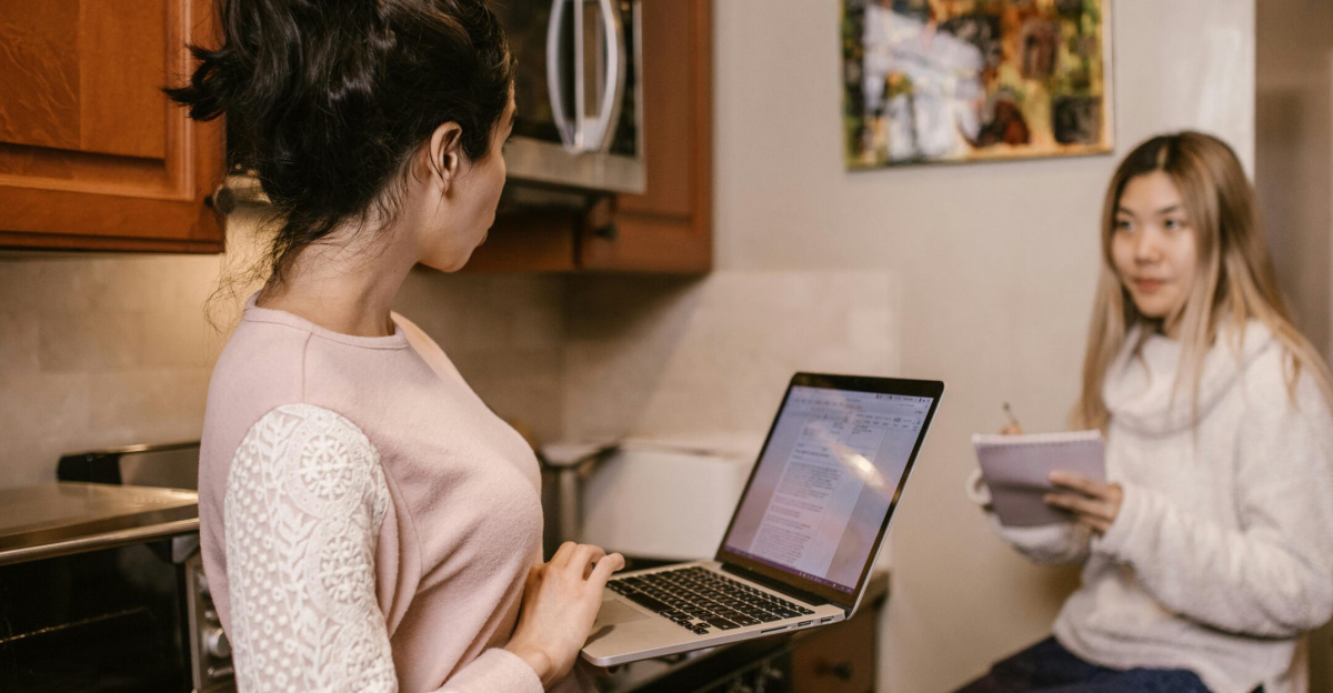 Two women collaborate on laptops and notes in a cozy home kitchen setting.
