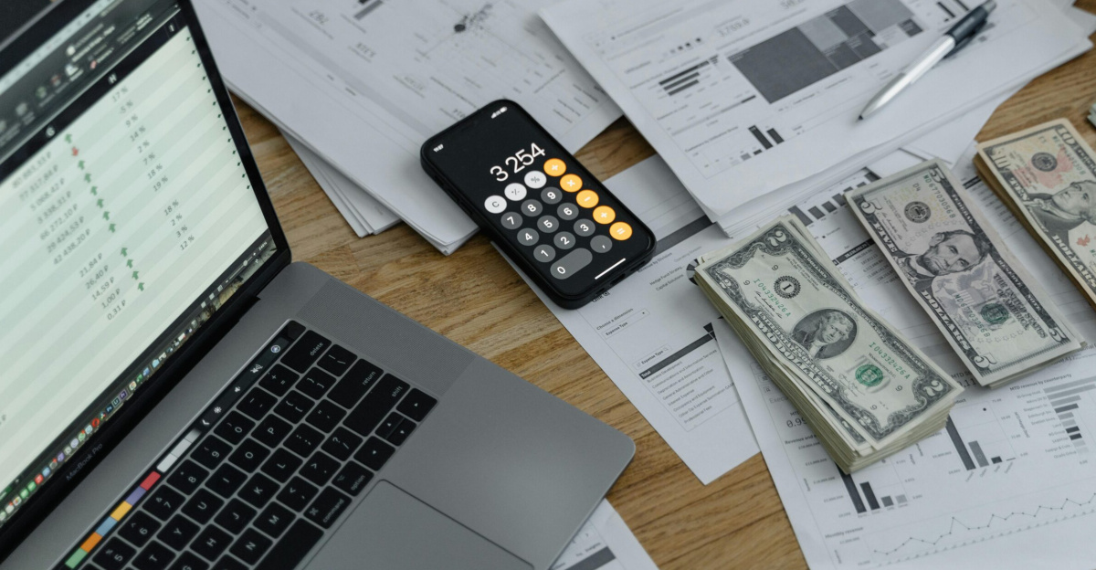 A desk with financial documents, currency, a laptop, and phone calculator.