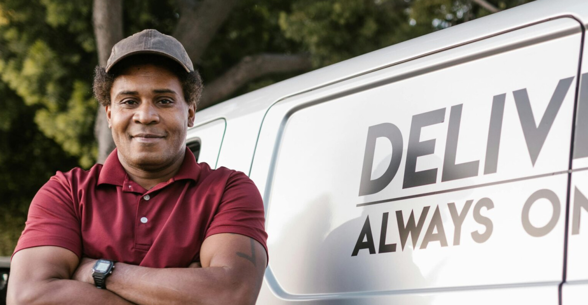 Delivery driver standing with arms crossed near a company van displaying a timely service slogan.
