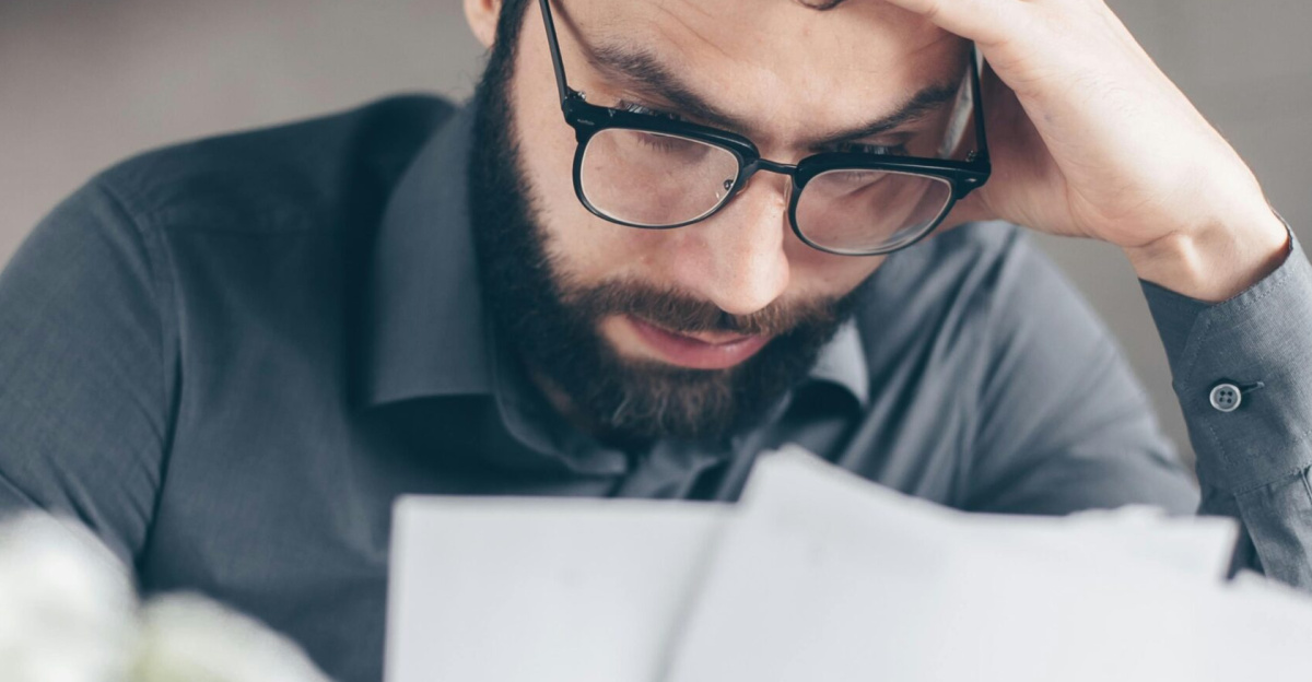 A worried man with glasses reviews financial papers, highlighting stress and financial concern.