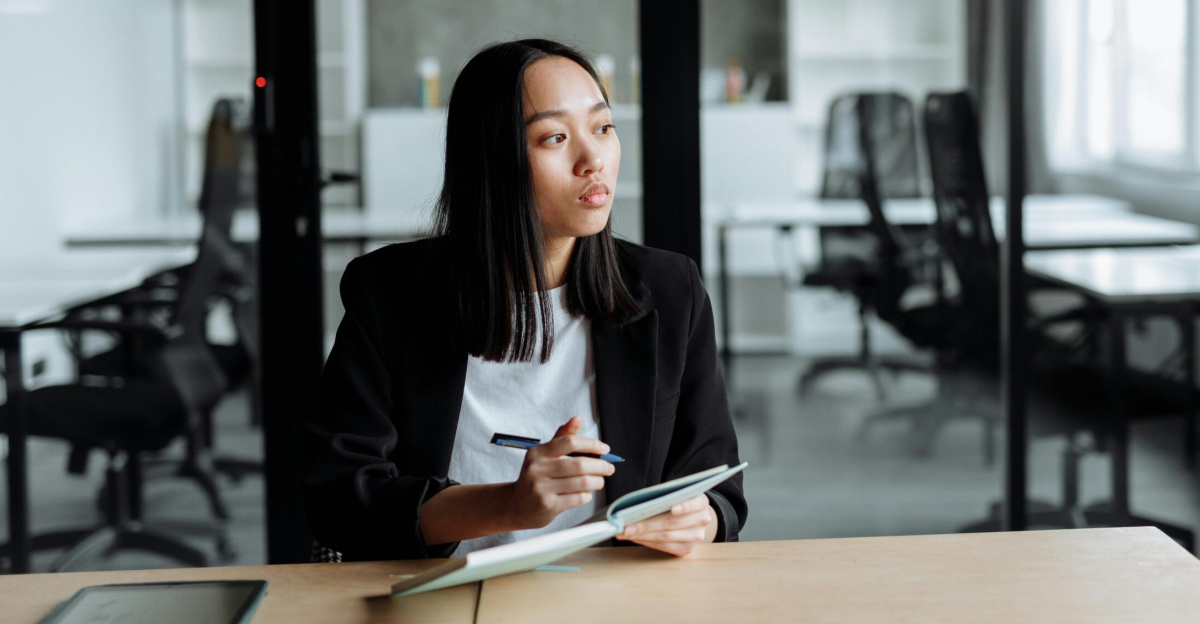 Portrait of a woman in a modern office pondering ideas, holding a notebook and pen.