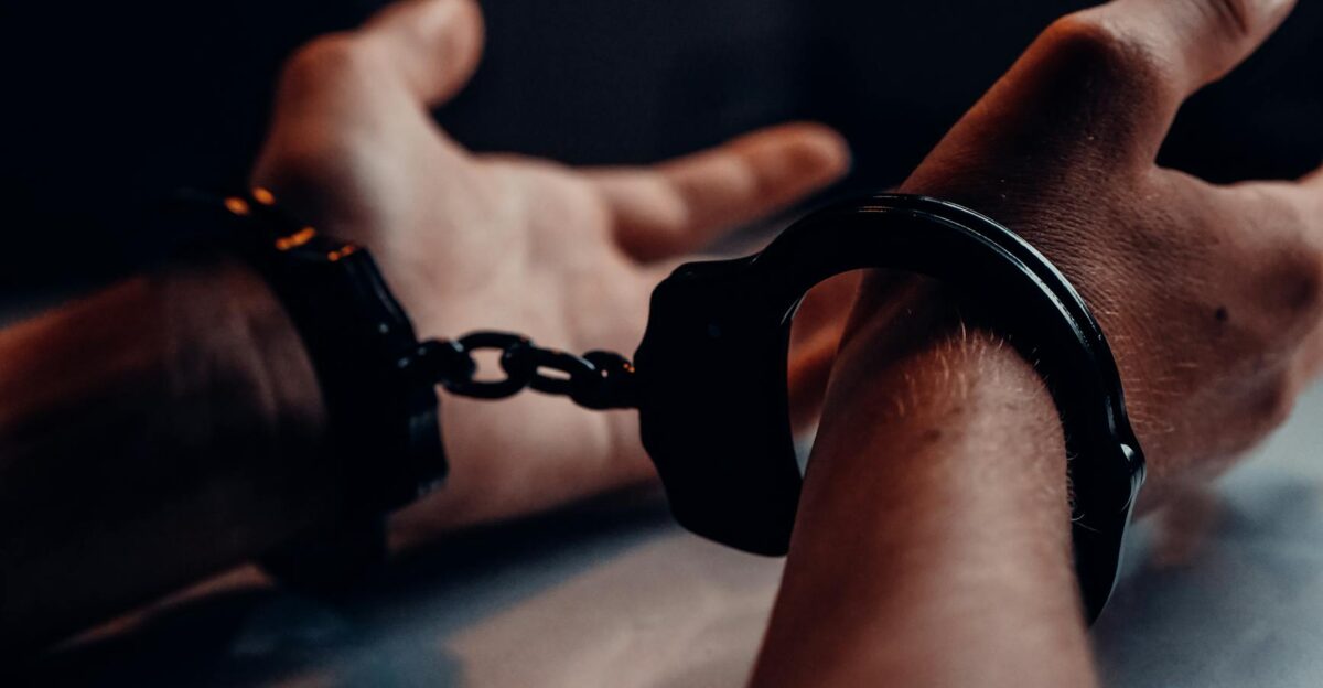 A close-up of human hands handcuffed on a table with a dark background depicting law enforcement themes