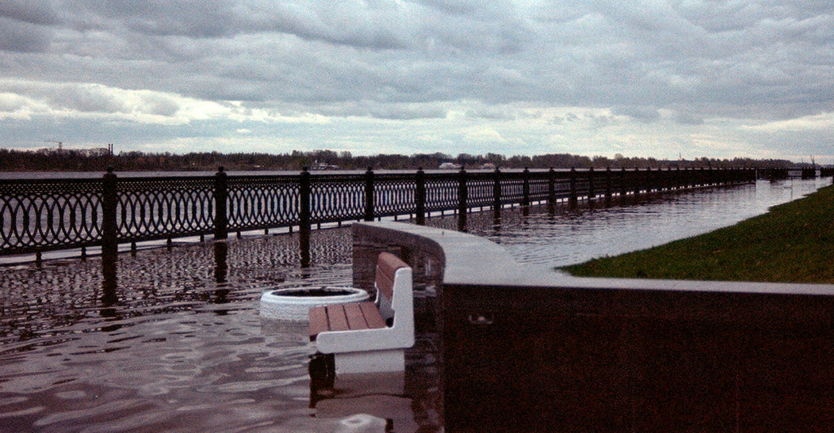 A flooded walkway by the river with overcast skies and empty benches submerged.