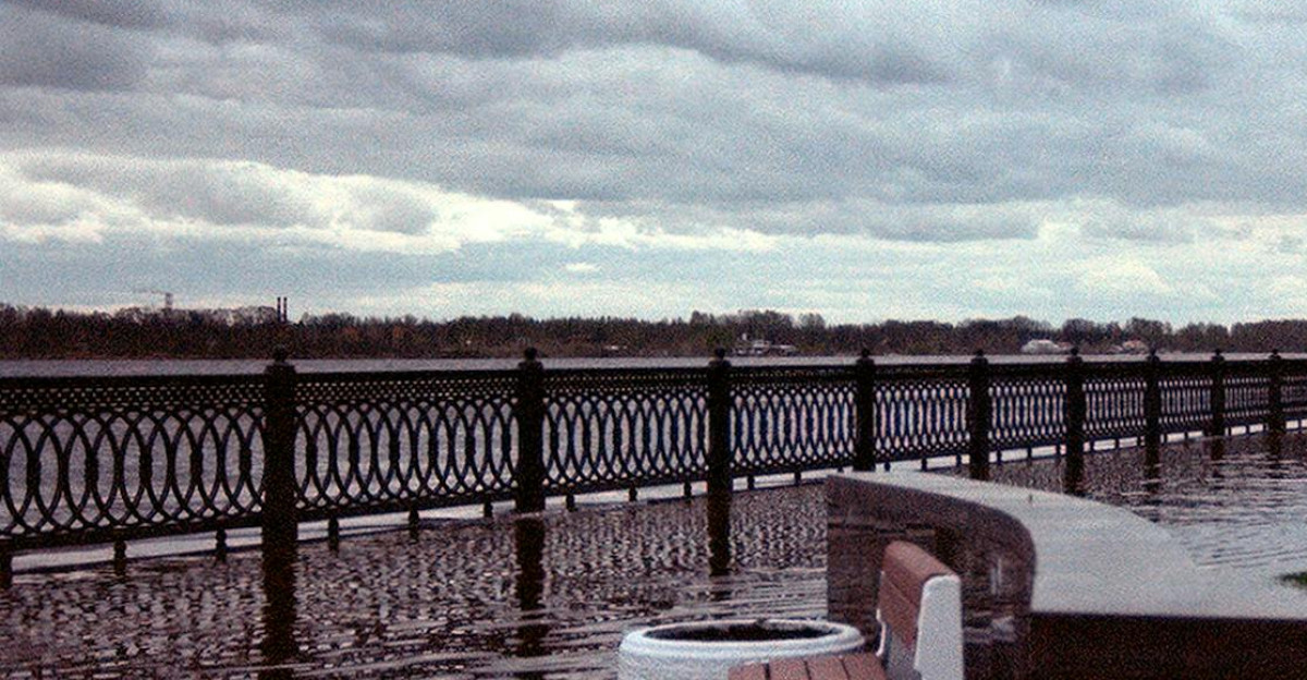 A flooded walkway by the river with overcast skies and empty benches submerged