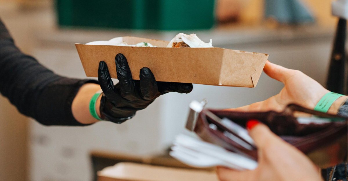 Person hands over food order in cardboard tray wearing black gloves in restaurant.