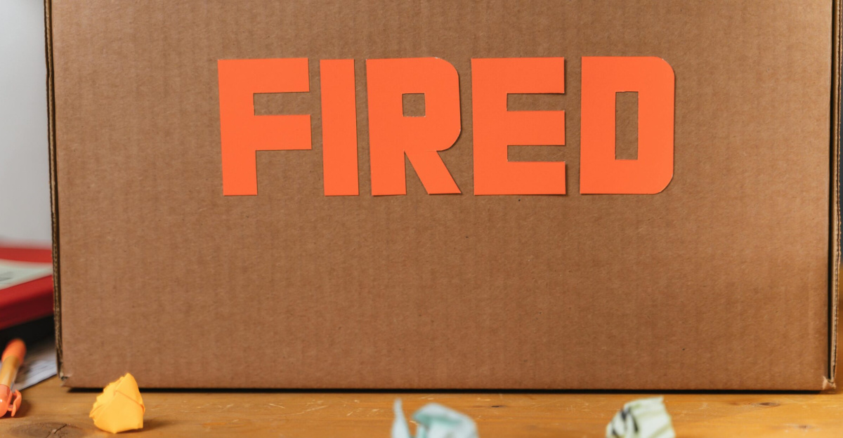 Close-up of a cardboard box labeled FIRED on a wooden table with crumpled papers.