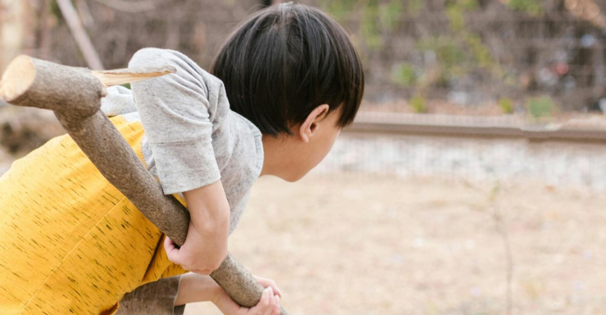 A child in a yellow shirt playing outside with a large tree branch on a sunny day.