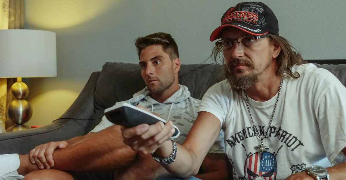 Two men relaxing on a sofa at home watching TV with a remote control in hand